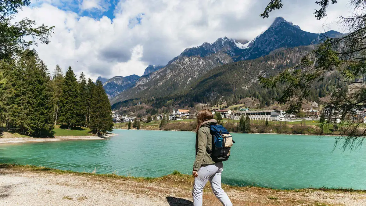 A hiker with a backpack on the pale shore of a green mountain lake under wooded slopes