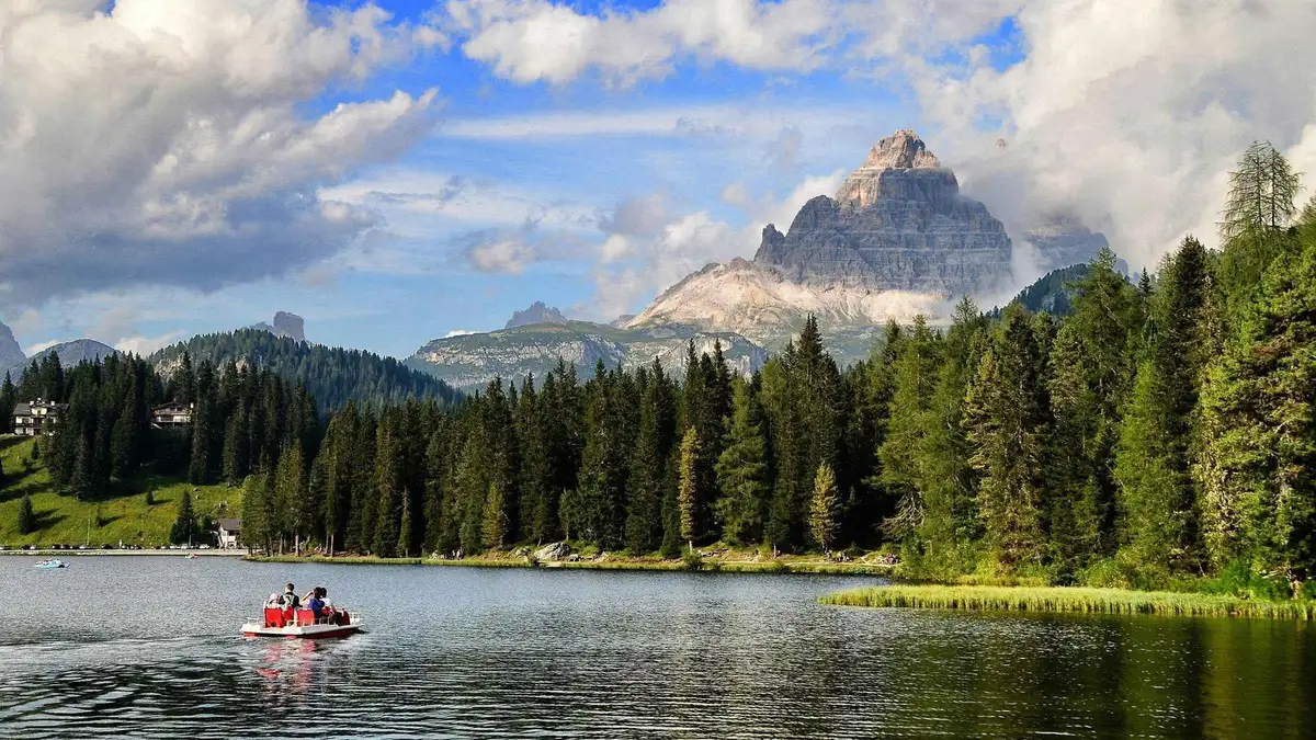 A pedal boat on dark green water below a lone dolomite tower and pine forest